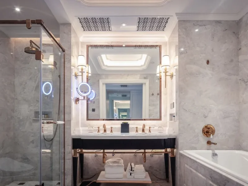 High-end luxury bathroom renovation featuring premium grey marble wall cladding, a grand backlit mirror with a gold frame, polished gold faucets, a glass-enclosed shower, and an integrated bathtub.