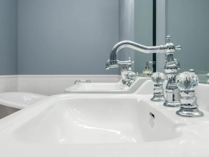 High-end traditional bathroom renovation showcasing a close-up of a double vanity with twin white ceramic sinks, elegant high-arc chrome faucets with crystal knobs, and soft blue-grey walls with white wainscoting.
