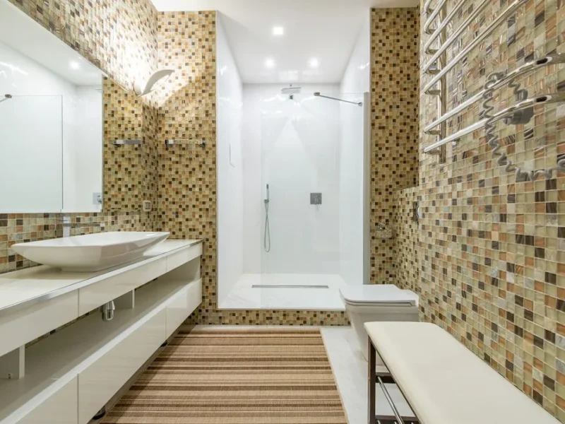 Modern luxury bathroom renovation featuring Earth-toned glass mosaic wall tiling, a long floating white vanity with a vessel sink, a large frameless mirror, and a seamless walk-in shower.