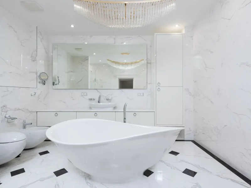 High-end luxury bathroom renovation featuring seamless white marble walls and floor, a modern freestanding slipper bathtub, a large vanity mirror, and a crystal chandelier.