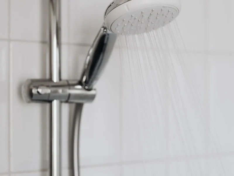 Modern chrome handheld shower head with water running, mounted on a slider rail against a background of clean white ceramic wall tiling.