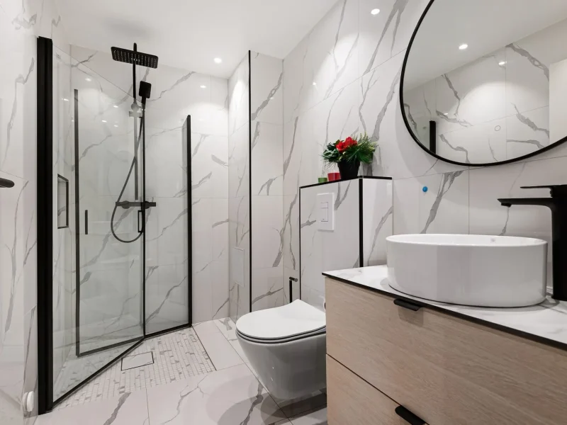 Modern luxury bathroom renovation featuring white marble wall tiles, a walk-in glass shower with matte black trim, a floating wood vanity with a vessel sink, and a round black-framed mirror.