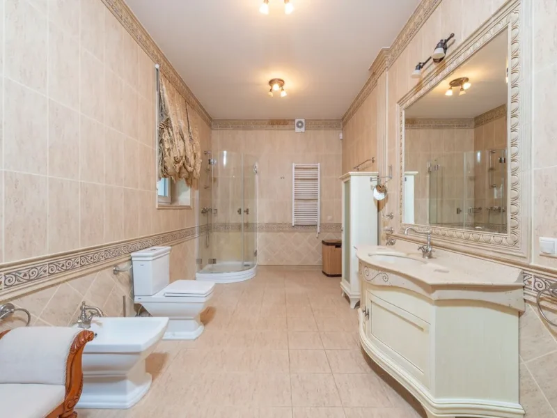 Spacious traditional luxury bathroom renovation featuring floor-to-ceiling beige marble tiling, a classic curved vanity unit with a large mirror, a white toilet and bidet set, and a curved glass corner shower.