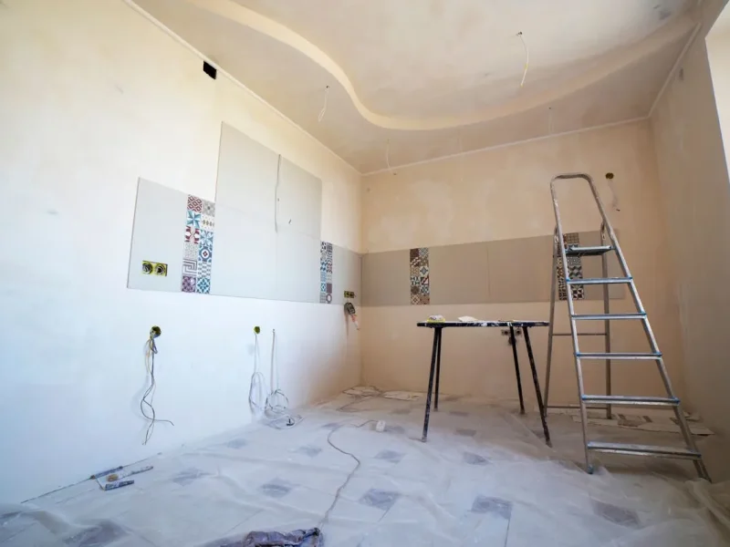 Residential room undergoing a professional renovation with freshly installed wall tiles featuring patterned accents, raw plastered walls, exposed electrical wiring, an aluminum stepladder, and a work table.
